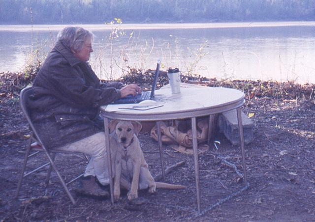 Cheryl Munson recording data at a table with a dog under the table on the river bank