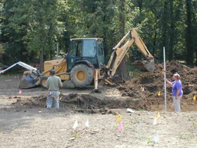 A backhoe removing soil while two people guide the mechanical excavation