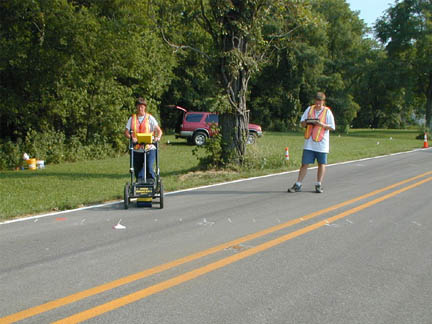 Two people in orange vests, one pushing the GPR unit over the paved road.