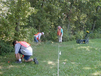 Three people in orange vests measure and flag the corners of a survey grid